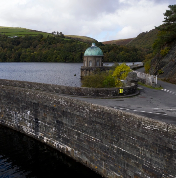 llun o'r afon yn cael ei dal yn ôl gan yr argae gyda choedwig a mynyddoedd yn y cefndir