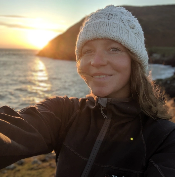 Dynes yn tynnu llun ohoni ei hun gyda'r môr a machlud haul yn y cefndir