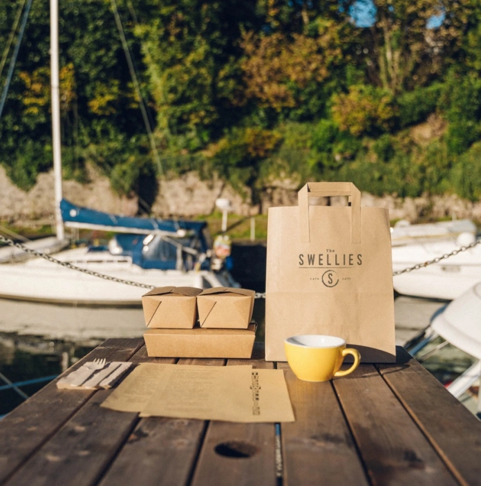 Bag papur brown gyda logo Swellies, tri blwch bwyd a mwg coffi melyn ar fwrdd pren gyda chwch ar y dŵr yn y cefndir
