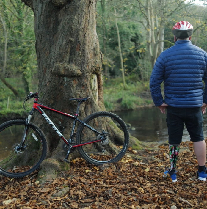 beic mynydd yn pwyso yn erbyn coeden mewn parc a pherson â thrwyn prosthetig yn sefyll wrth ei ochr.