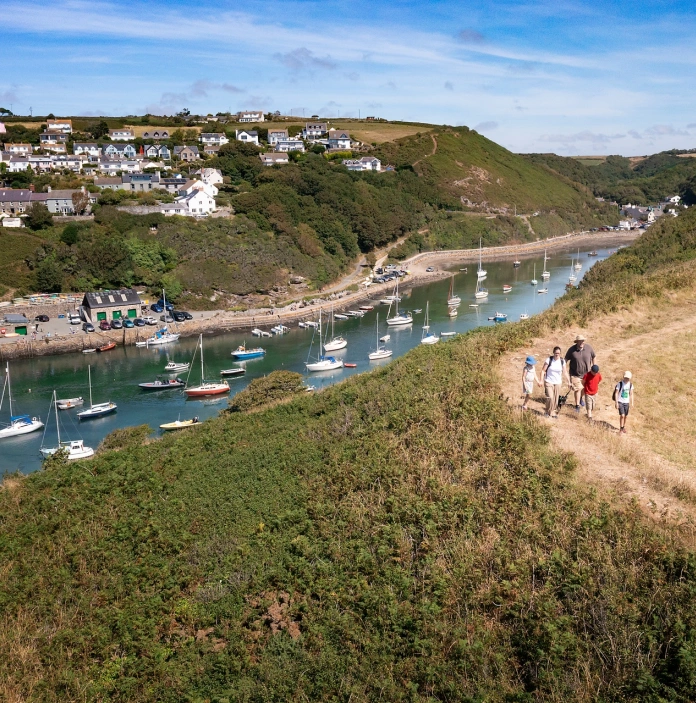 A harbour village surrounding a boat-filled estuary - houses one side, walkers on a path on the other.