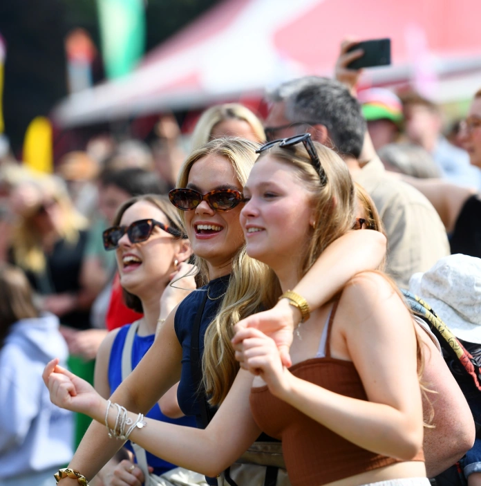 Three friends with arms around each other singing along as part of a festival crowd.