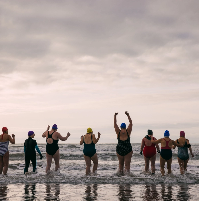ten individuals going cold water swimming wearing swimsuits and swim caps. Seen from behind, they are stood in the shallow waves of the sea. One person is holding their hands above their head, others have their hands in the air.