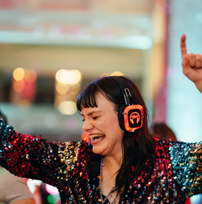 a person is a rainbow coloured glittery top wearing headphone and dancing with their hands in the air.