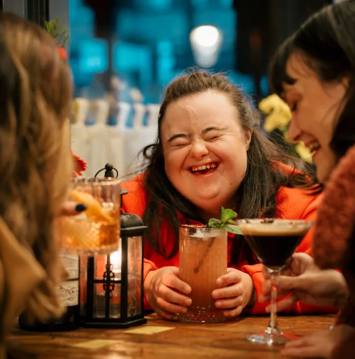 Three people sat around a table with cocktails, the central person is laughing with eyes closed and mouth open.