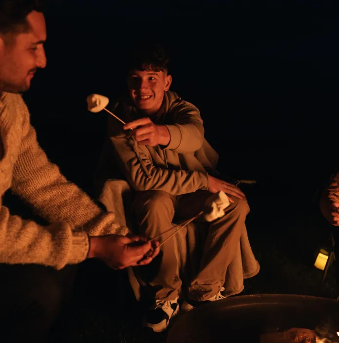 A adult and two teenage children sat by a fire toasting marshmallows,