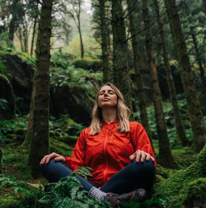 A person sat crossed legged with eyes closed in a forest, relaxing.
