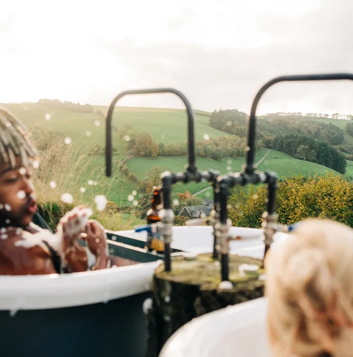 two people sat side by side in individual outdoor bathtubs blowing bubbles from the bath at each other and laughing