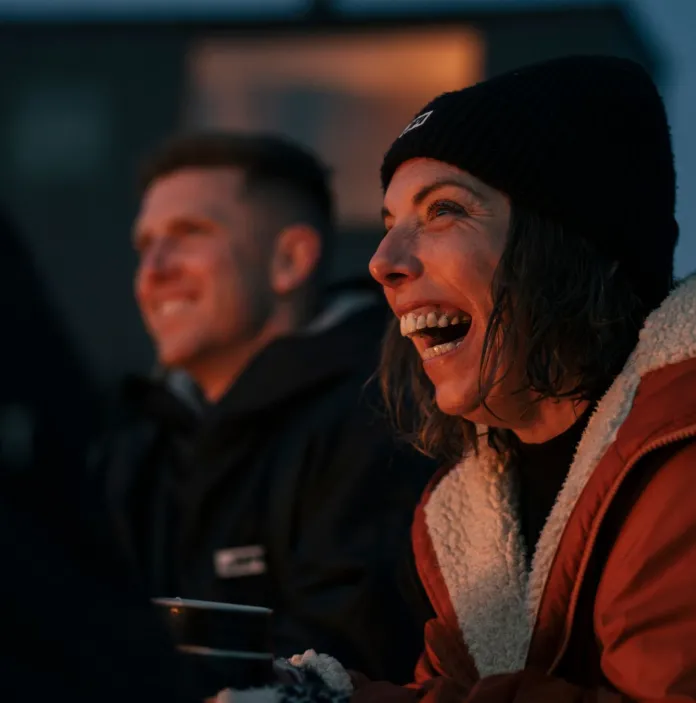 two people sat together at dusk, their smiling faces highlighted by light from a fire.