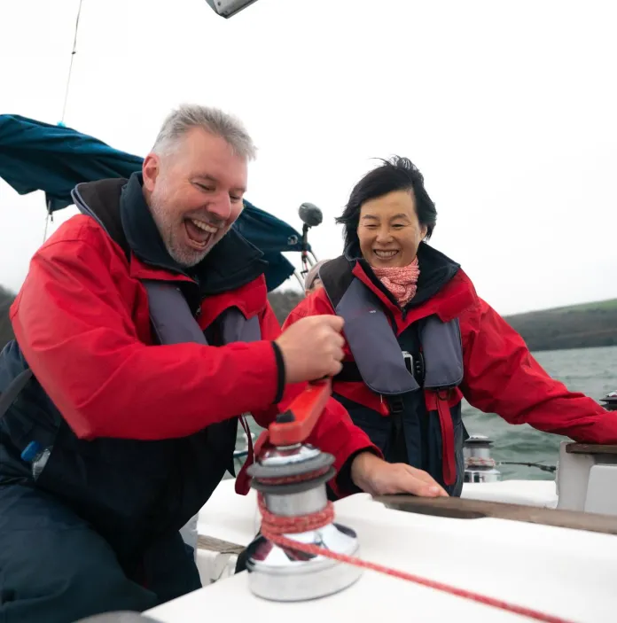 two people on a boat wearing red and grey coats, with wind swept hair and big smiles. The peron in the foreground is wind in the sail reel.