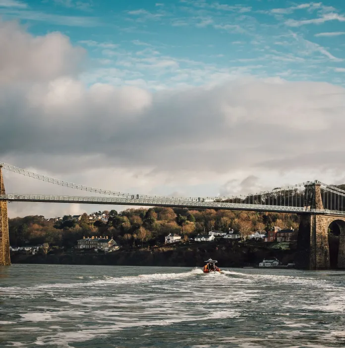 Llun o'r bont grog gyda choed a thai yn y cefndir a chwch ar Afon Menai