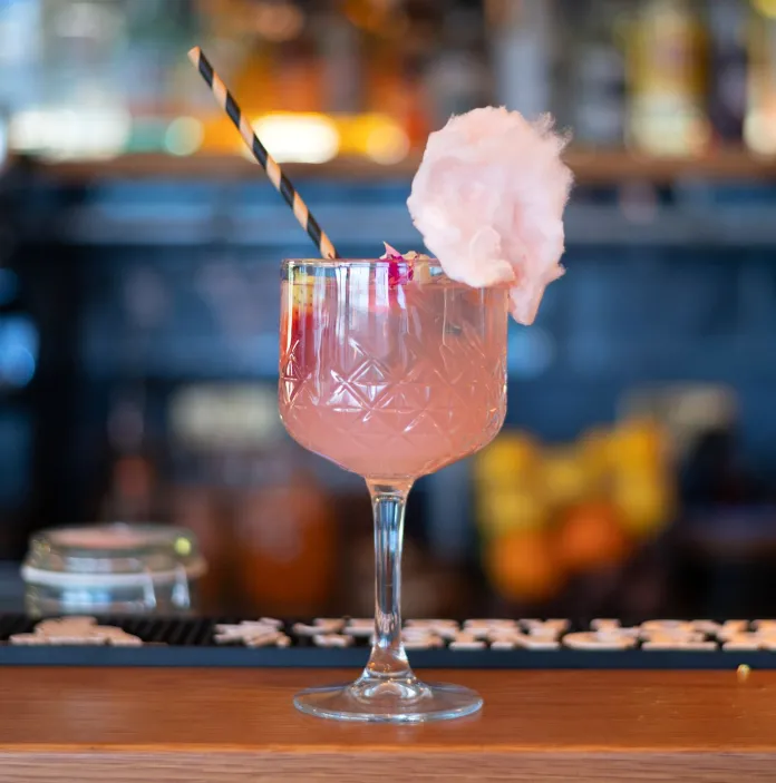 Mae coctel pinc mewn goblet gwydr yn eistedd ar flaen bar. Mae gan y coctel wellt streipiog a darn o candyfloss ar ochr y gwydr.