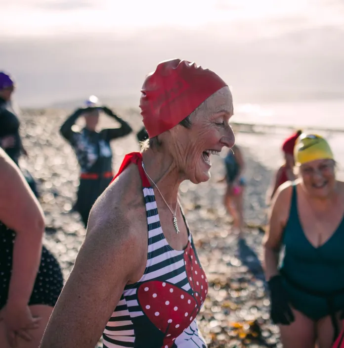 Aelod o’r grwp nofio ‘the blue tits’ yn sblasio yn y môr yn Ynyslas gan chwerthin gyda ffrindiau