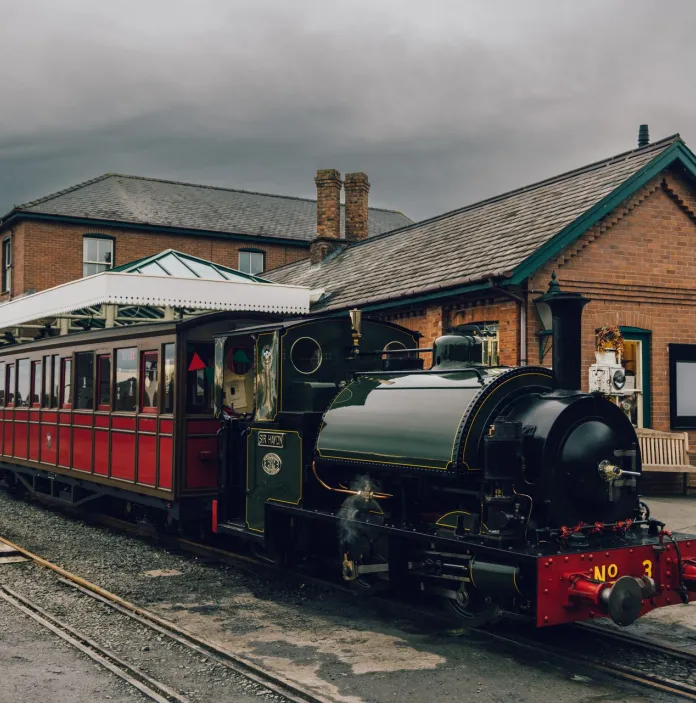 A small green steam loco and train at a narrow gauge railway station.