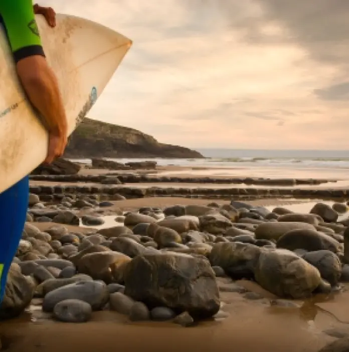 Llun o berson gyda'u cefn at y camera, yn wynebu'r traeth a'r môr, gyda bwrdd syrffio dan eu cesail