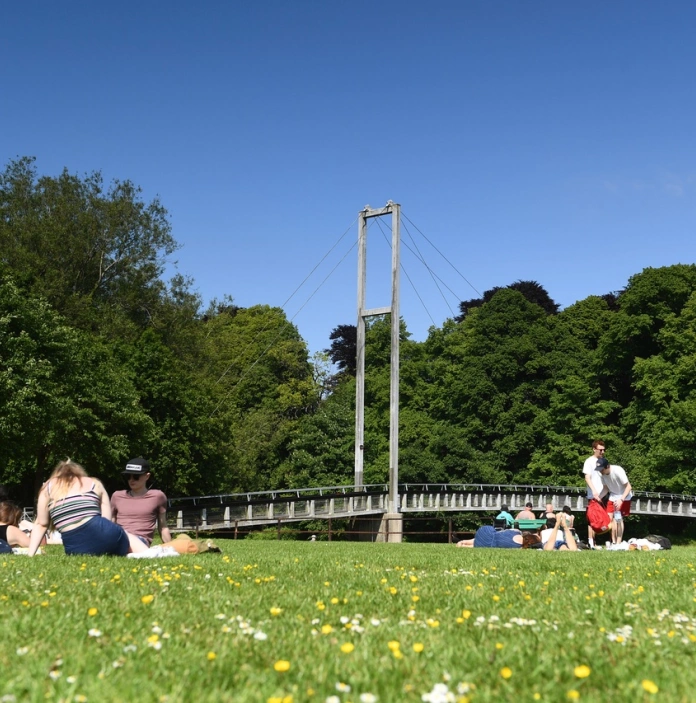 Pobl yn eistedd mewn parc ger Pont Blackweir yng Nghaerdydd ac awyr las