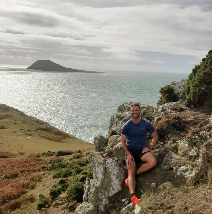 Dyn yn eistedd ar graig gydag Ynys Enlli yn y cefndir