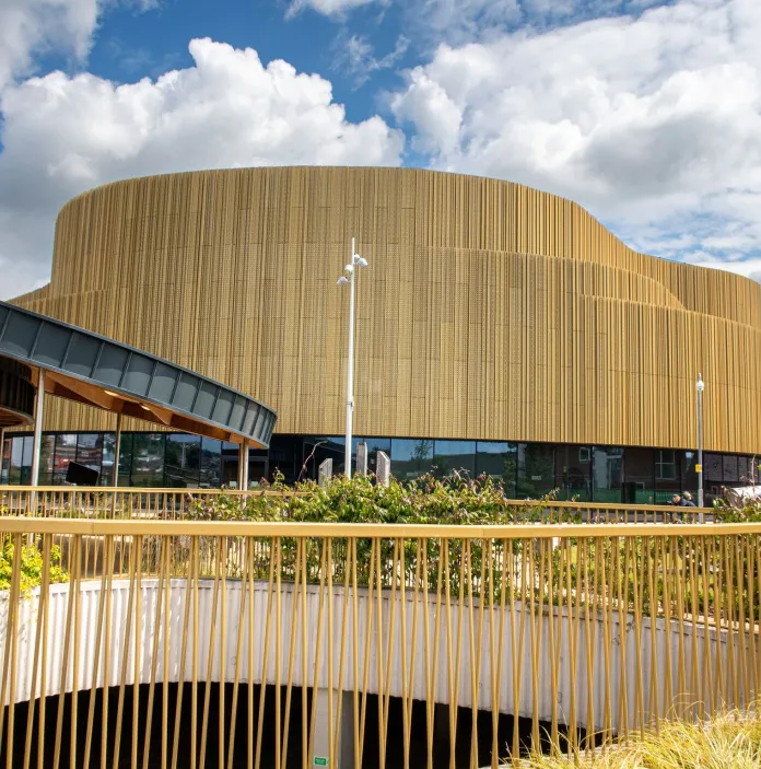 Tu allan i arena Abertawe, adeilad gyda waliau pren a gwyrddni o'i flaen.