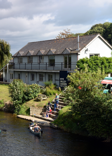 Pobl yn canwio ar yr Afon Gwy gydag adeilad Wye Valley Canoes ar y lan yn y cefndir