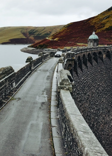 Car yn dod ar y ffordd hir dros bont garreg, gyda chronfa ddŵr i'r chwith, a bryniau yn y cefndir