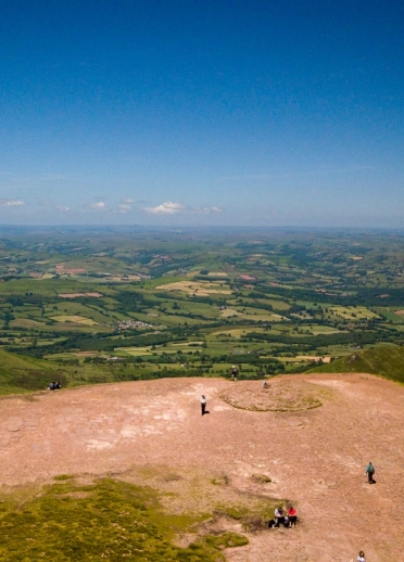 Golygfa o'r awyr o'r gwastadeddau gwyrdd islaw o ben Pen y Fan gydag awyr las.