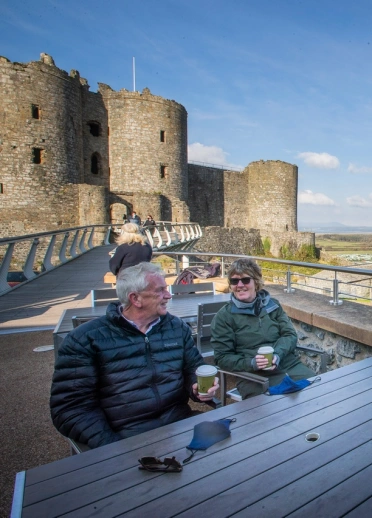 Cwpl hŷn yn yfed coffi i fynd y tu allan i gastell, yn mwynhau’r golygfeydd.