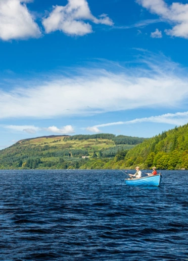 Llyn a bryniau yn y cefndir. Mae adeilad cronfa ddŵr ar y dde, a chwch bach glas gyda dau berson ynddi.