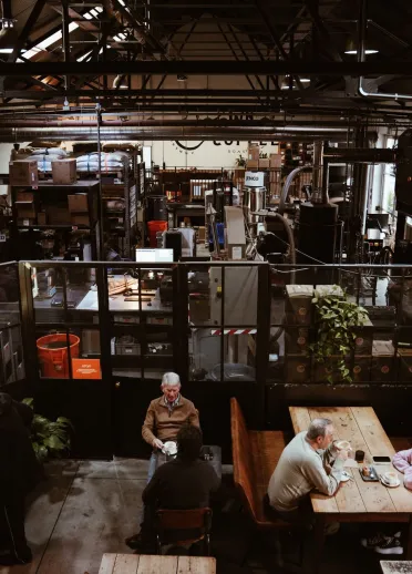 Here’s concise alt text for the image, with **no menti industrial-style café with customers sitting at wooden tables in the foreground and a large open roastery workspace visible behind glass panels.