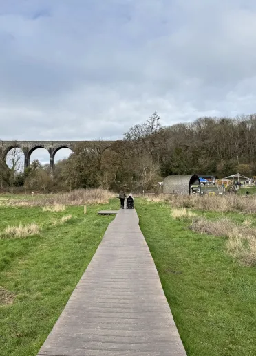 Llwybr pren mewn parc gwledig, gyda choed a thraphont reilffordd yn y cefndir.