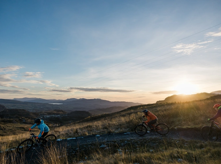 Tri person yn beicio mynydd i lawr trac ar ochr mynydd. Mae'r haul yn machlud tu ol i'r mynyddoedd.