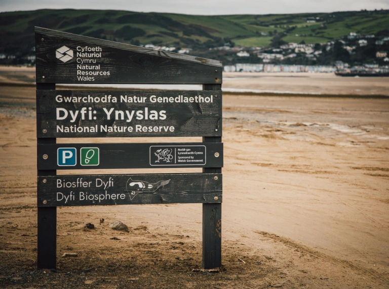 Arwydd gwarchodfa natur ar draeth tywodlyd gyda thref yn y cefndir pell.