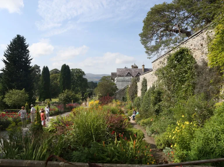 Flowers, trees and shrubs alongside paths and stone walls with a grand house in the background.