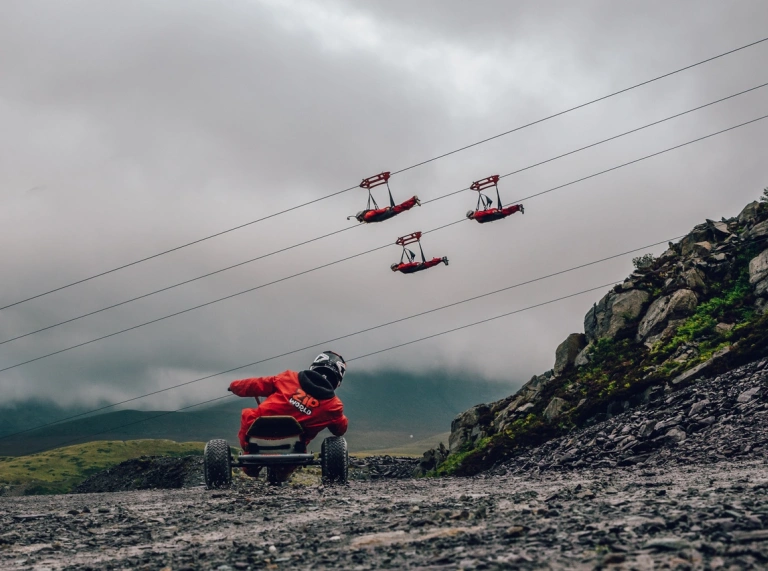 Gwib-gartiwr yn gyrru i lawr chwarel gyda phobl yn pasio heibio ar wifrau gwib.