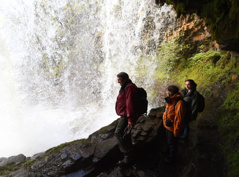 Tri person yn cerdded tu nôl i'r rhaeadr