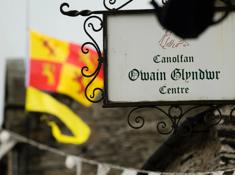 Llun o arwydd Canolfan Owain Glyndŵr, gyda baner Owain Glyndŵr yn chwifio yn y cefndir