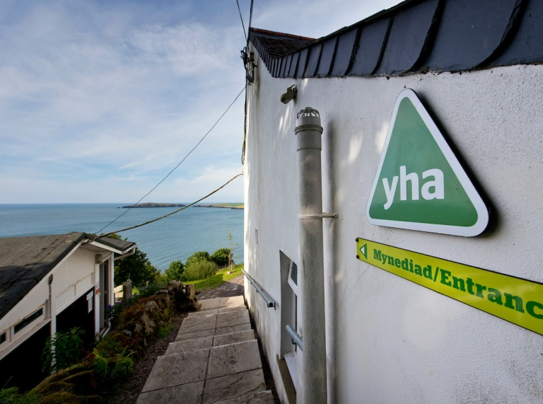 Mynediad i adeilad gwyn gyda’r môr yn gefndir.