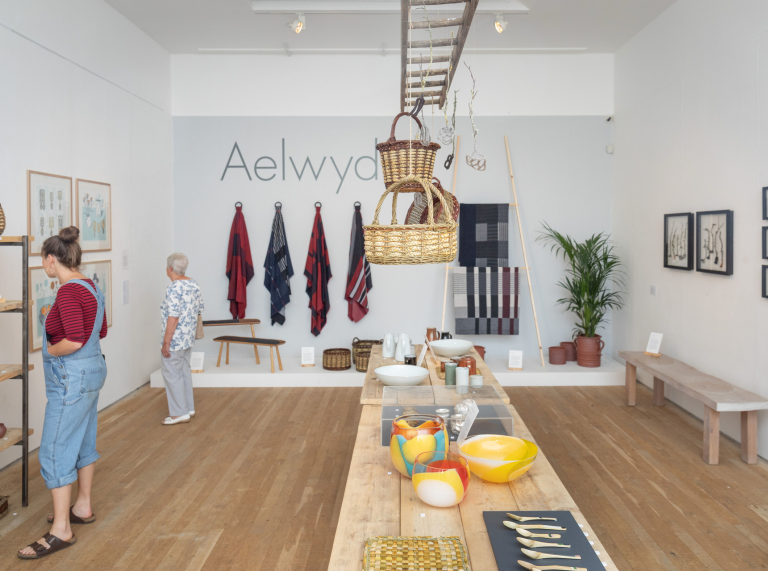 Crefftau’n cael eu harddangos ar fwrdd a wal, gyda dwy fenyw’n edrych ar waith celf ar wal yng Nghanolfan Grefft Rhuthun.