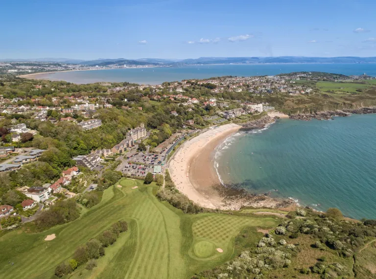 Llun o’r awyr o gwrs golff ac arfordir yn yr heulwen.