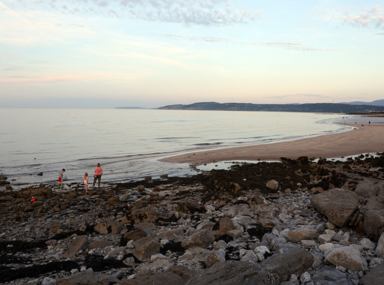 Teulu yn archwilio traeth creigiog a thywod yn y pen pellaf