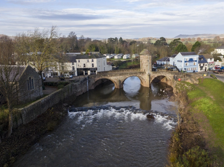 Pont gerrig hanesyddol yn croesi afon, gyda thai a siopau ar hyd glannau’r afon a chored yn y blaendir.