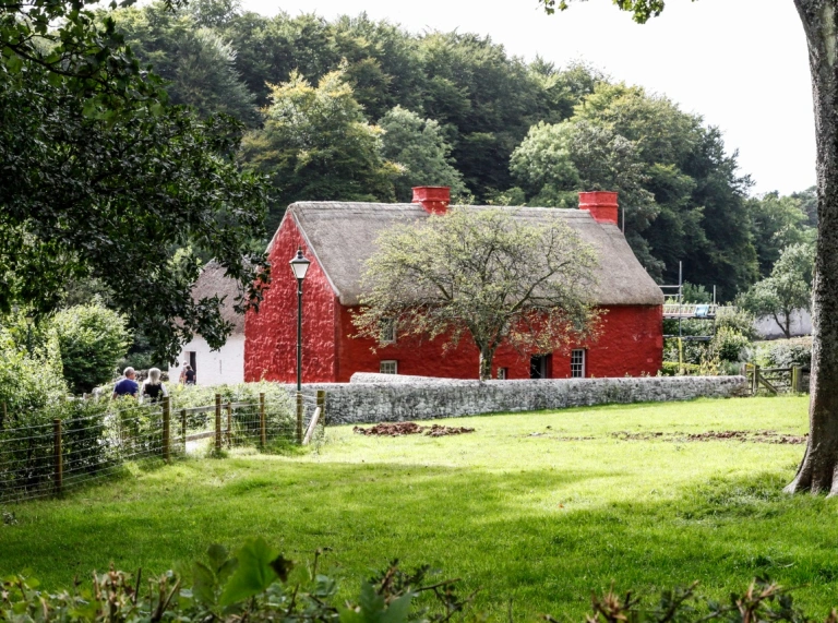 Bwthyn coch wrth ymyl cae gyda choed y tu ôl iddo
