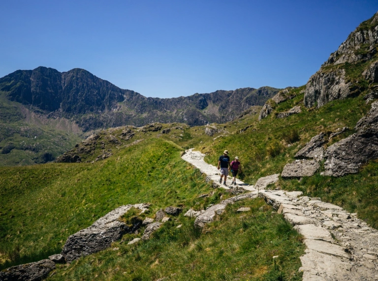Dau berson yn heicio i fyny llwybr mynydd gydag awyr las
