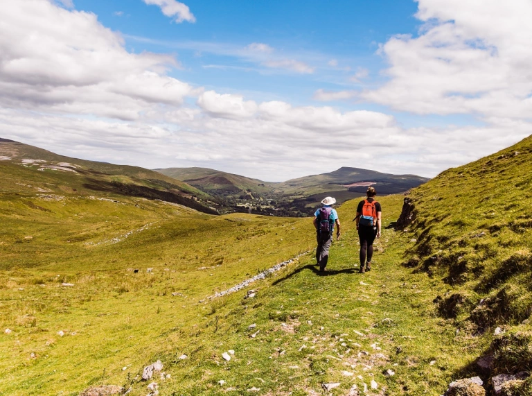 Dau gerddwr ar lwybr sy’n arwain at fynyddoedd dramatig.