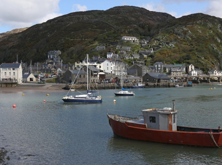 Delwedd o gychod bach yn harbwr Barmouth a thai a bryniau yn y cefndir.
