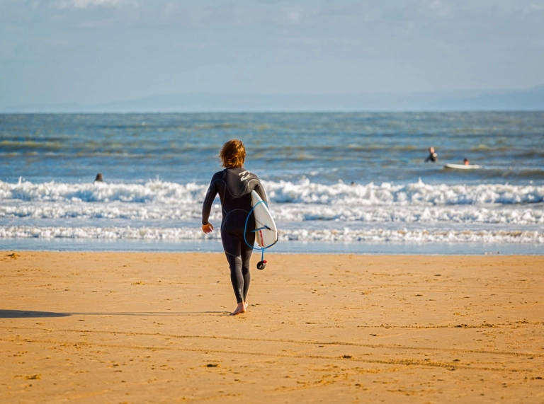 Person yn cerdded ar y traeth tuag at y môr gyda bwrdd syrffio ar draeth Porthcawl