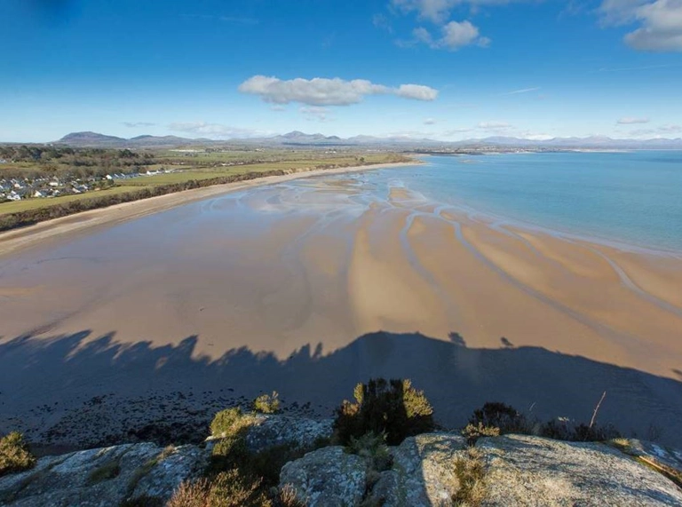 traeth tywodlyd eang gydag arfordir a bryniau yn y pellter.
