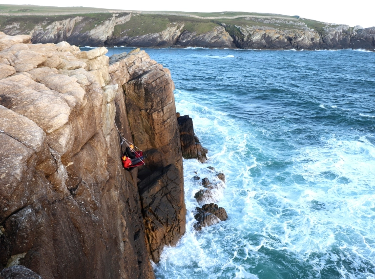 Wyneb clogwyn uwchben y môr gyda dau berson yn eistedd ar ‘portaledge’ sy’n hongian o ymyl y clogwyn.