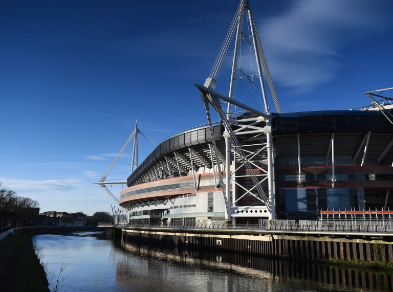 Stadiwm Principality Caerdydd o lan yr afon, gydag adlewyrchiad o’i mastiau trawiadol yn y dŵr o dan awyr las glir.