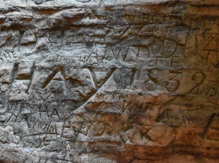 Enwau a rhifau wedi’u cerfio ar graig mewn ogof.