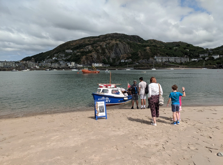 Grŵp o bobl yn sefyll ger fferi fach ar draeth tywodlyd.